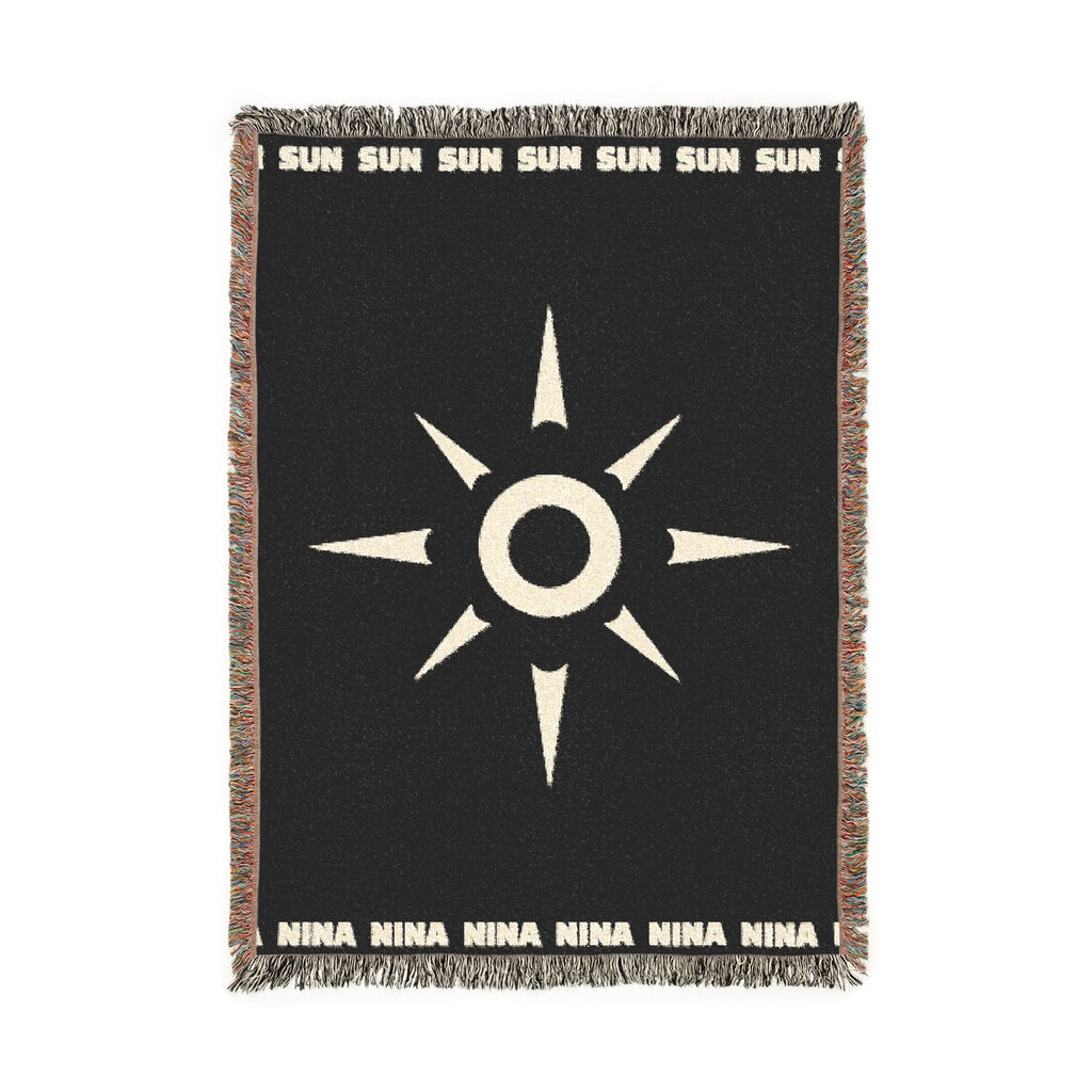 A black woven throw blanket with a central white SUN NINA sunburst emblem, white text at the top and bottom, and multicolored fringed edges, shown flat against a white background.